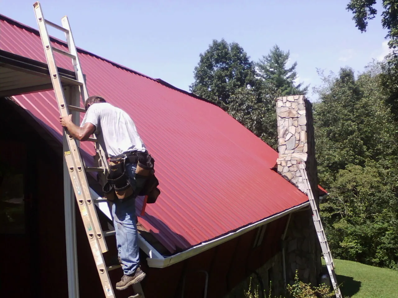Roofer on a ladder beside a red metal roof and stone chimney — Soffit & Fascia Repair by Roofing Brothers in Johnsonville